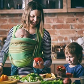 A mother preparing food with baby in a sling and a toddler